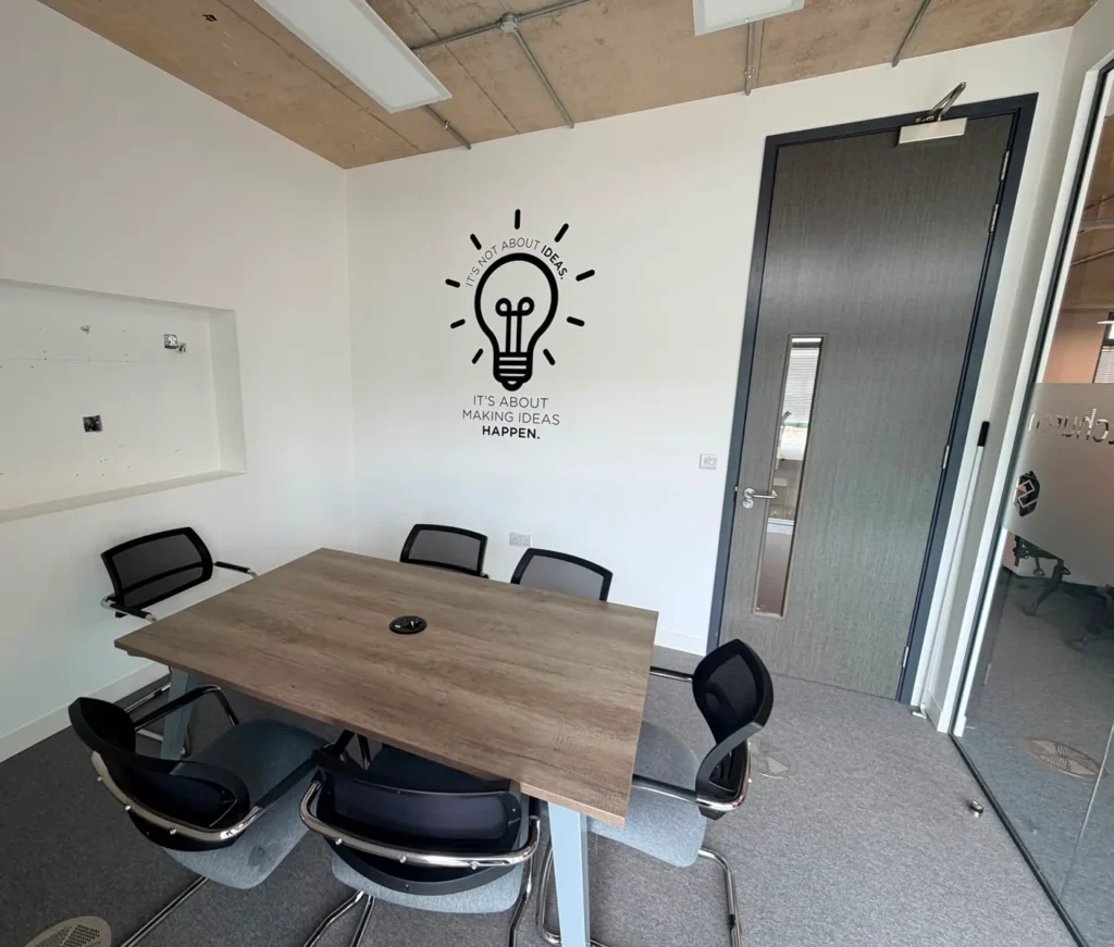 A small, professional meeting room featuring a wooden table and black chairs, accented by an inspiring "It’s not about ideas, it’s about making ideas happen" wall decal. This modern workspace is designed for collaborative team discussions and focused work sessions.