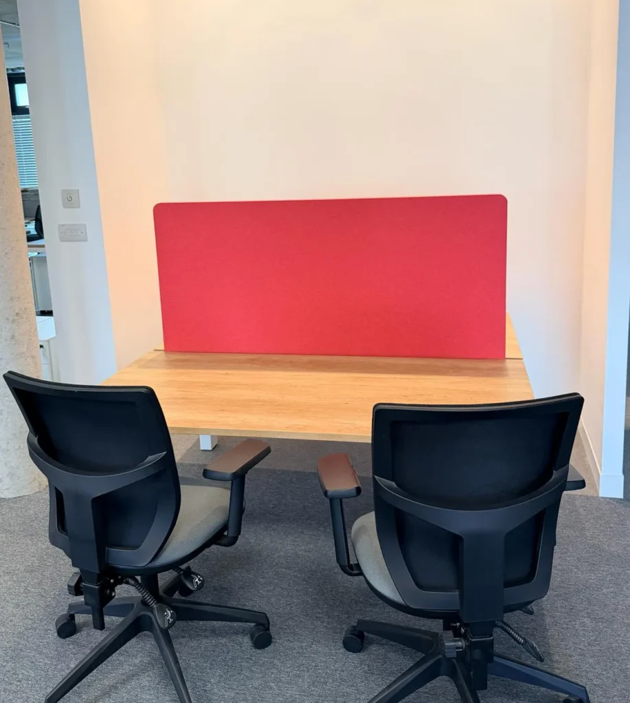 A clean, contemporary office workspace featuring a wooden desk, two ergonomic black office chairs, and a vibrant red acoustic privacy screen, designed for a professional meeting or focused work area.